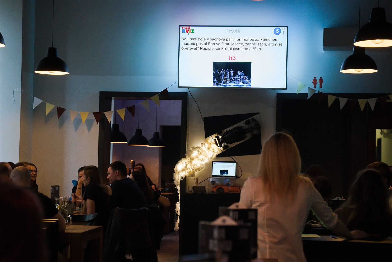 Kvízový večer v podniku – hráči pri stoloch a otázka na projekcii